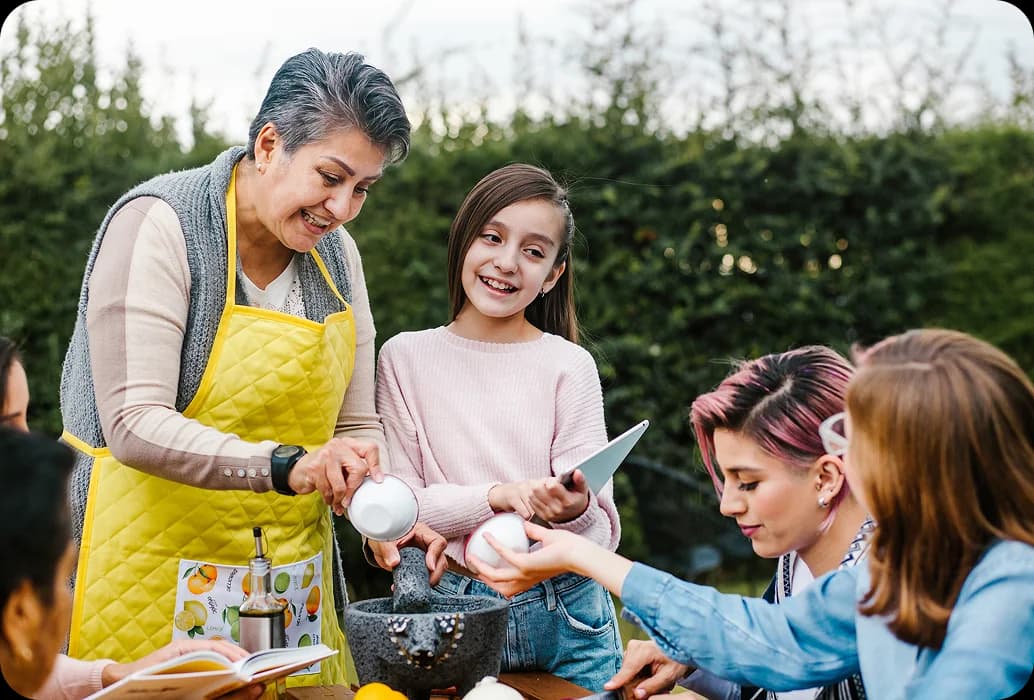 An adult teaches children to prepare ingredients with a mortar and pestle during a hands-on cultural learning activity outdoors.